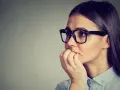 Zmartwiona kobieta w okularach obgryza paznokcie. Czy to objaw kleptomanii? Przyczyny tego zaburzenia są złożone.
