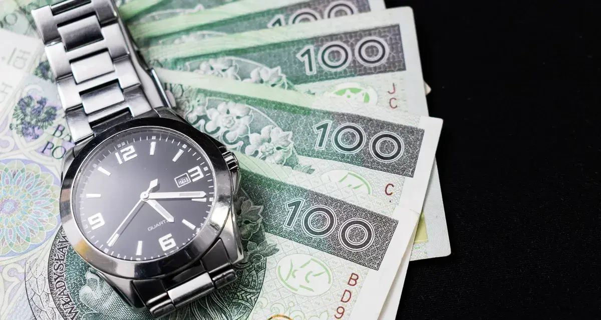 Zegarek na stosie banknotów 100 zł. Czas to pieniądz, a ten schemat zachowań kompulsywnych pokazuje, jak łatwo można stracić kontrolę nad finansami.