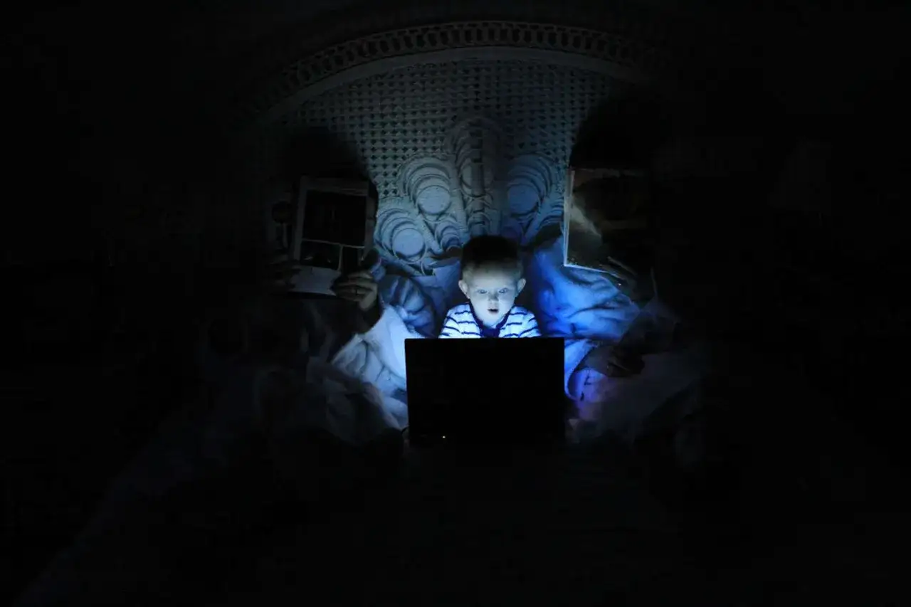 Dziecko wpatrzone w ekran laptopa, otoczone przez osoby czytające. Obrazek ilustruje problem siecioholizm definicja.