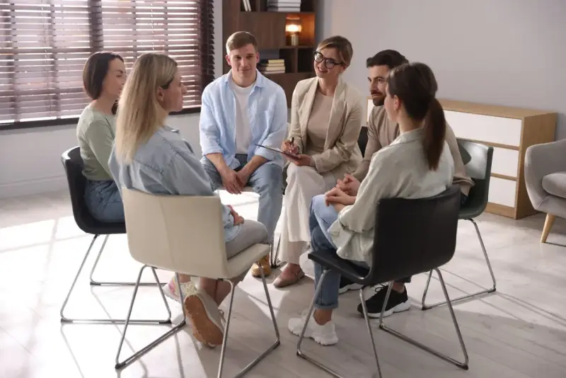 Grupa ludzi siedzi w kręgu, słuchając terapeuty. To scena z ośrodka dla uzależnionych, gdzie wsparcie i zrozumienie pomagają w powrocie do zdrowia.