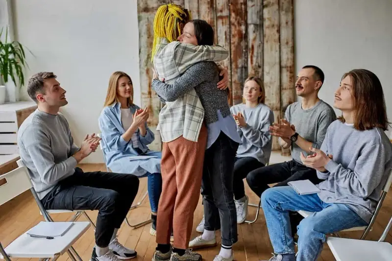 Grupa ludzi siedzi w kręgu, klaszcząc. Dwie osoby w centrum mocno się przytulają. Co to aa? Wsparcie i akceptacja.